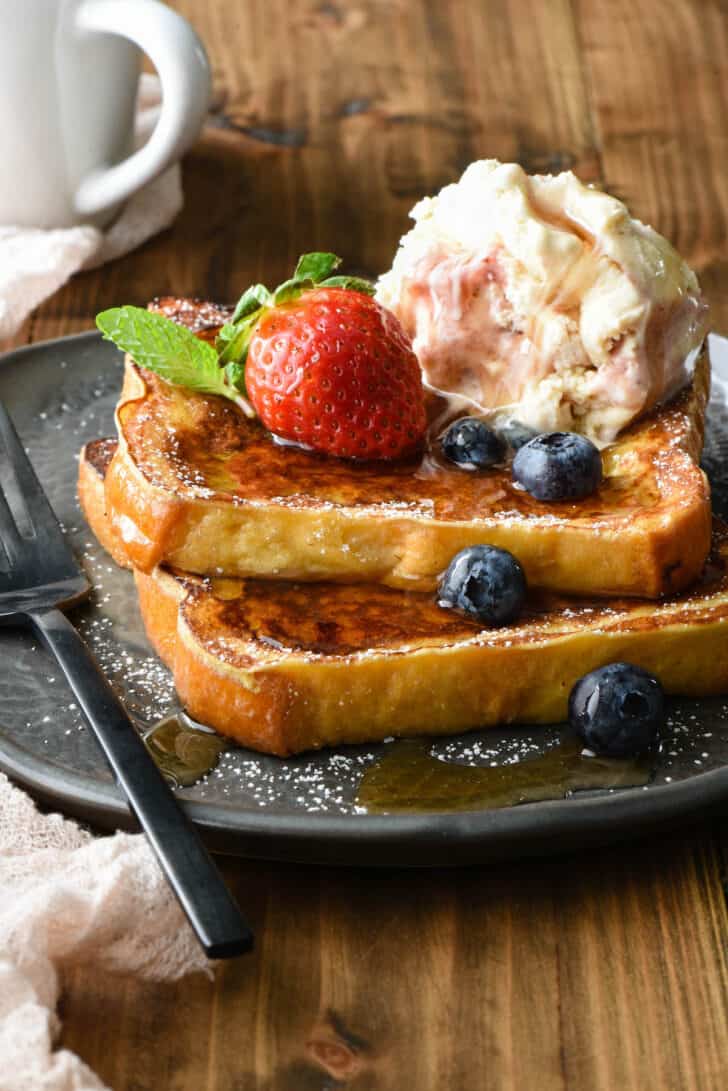Two slices of ice cream French toast on a gray plate, topped blueberries, mint, a strawberry, a scoop of ice cream and maple syrup.