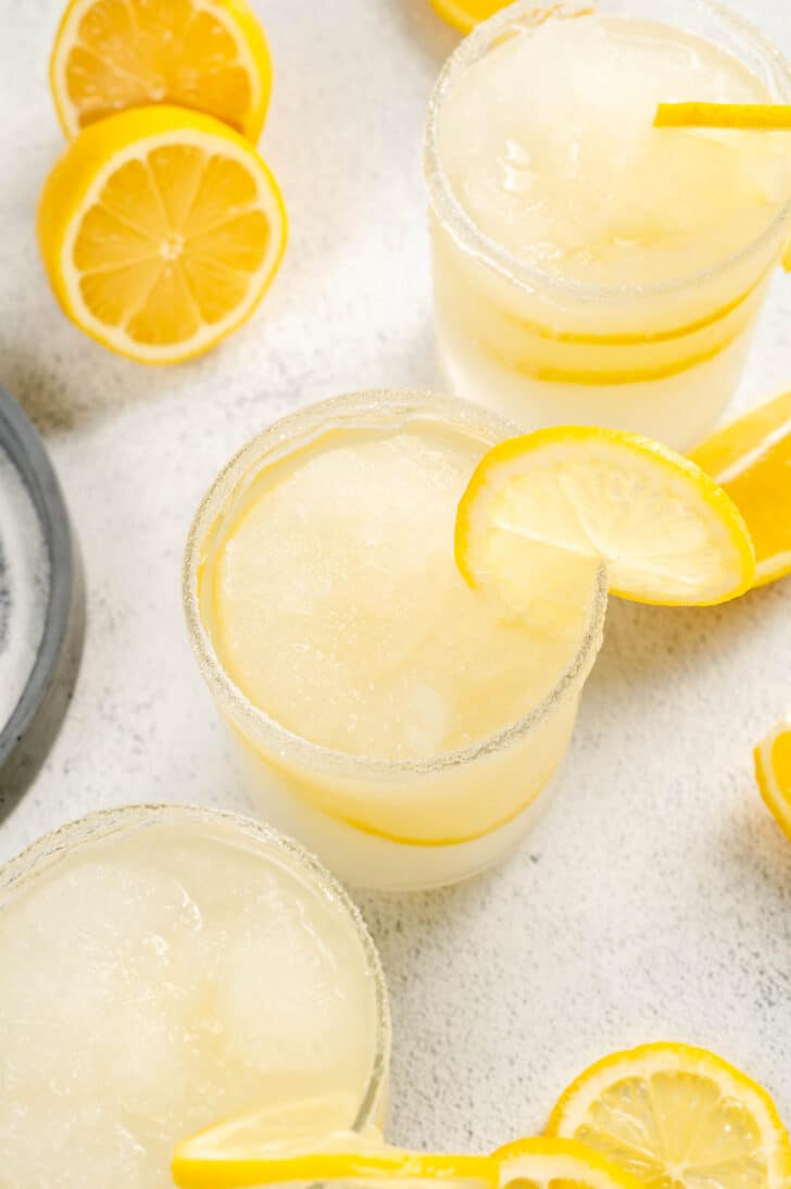 Three glasses of lemon margarita garnished with lemons slices, on a light surface.