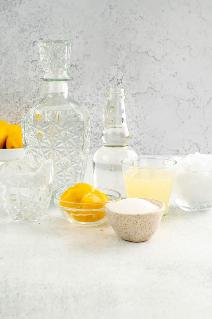 All of the ingredients needed for a margarita with lemon, including clear bottles of tequila and triple sec, a glass of lemon juice, lemon peels, sugar and ice.