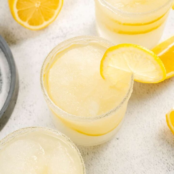 Three glasses of lemon margarita garnished with lemons slices, on a light surface.