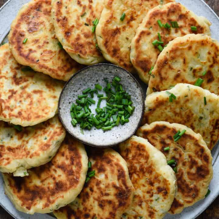 Gray plate topped with mashed potato cakes arranged in a circle, with a small bowl of chives in center of circle.