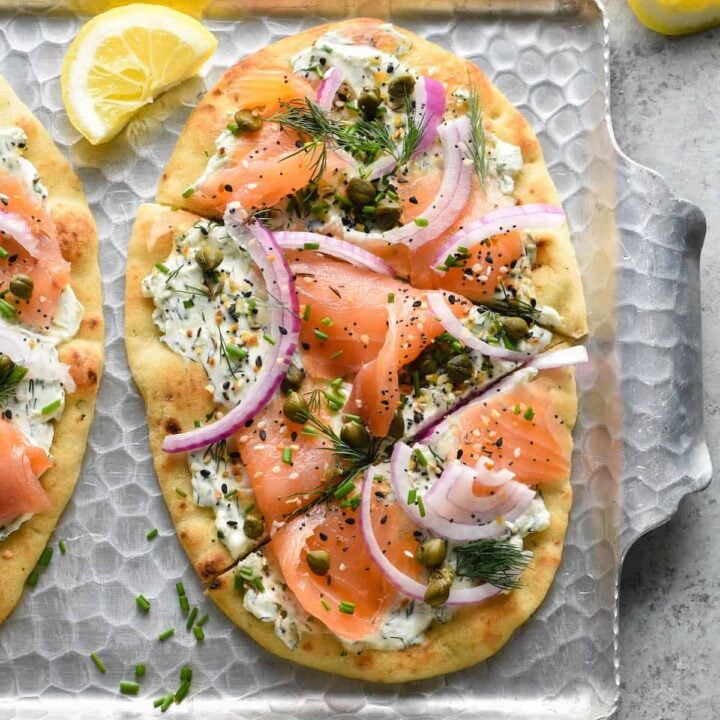 A smoked salmon pizza on a metal textured tray made with naan bread, cream cheese, herbs, everything bagel seasoning, lox, capers and red onion.