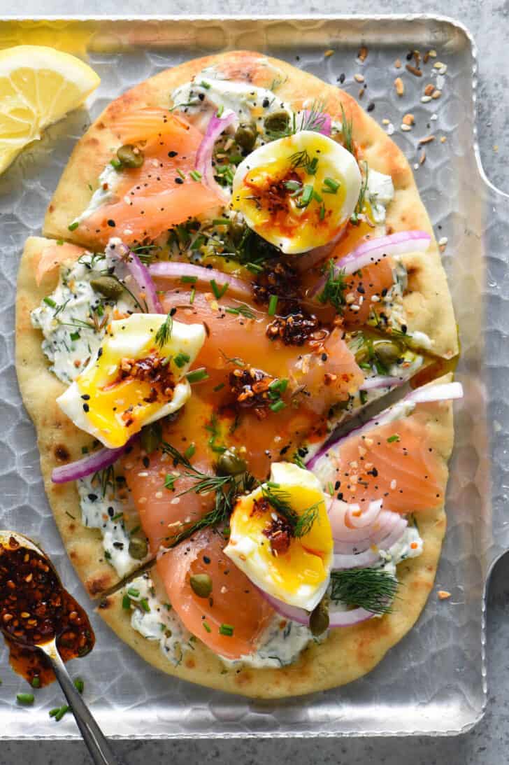 A smoked salmon pizza made with naan bread, herb creamed cheese, red onions, capers, soft eggs and chili crisp.
