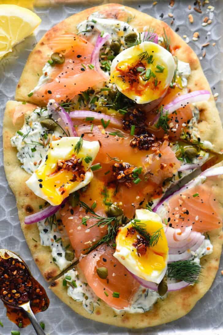 A smoked salmon pizza made with naan bread, herb creamed cheese, red onions, capers, soft eggs and chili crisp.