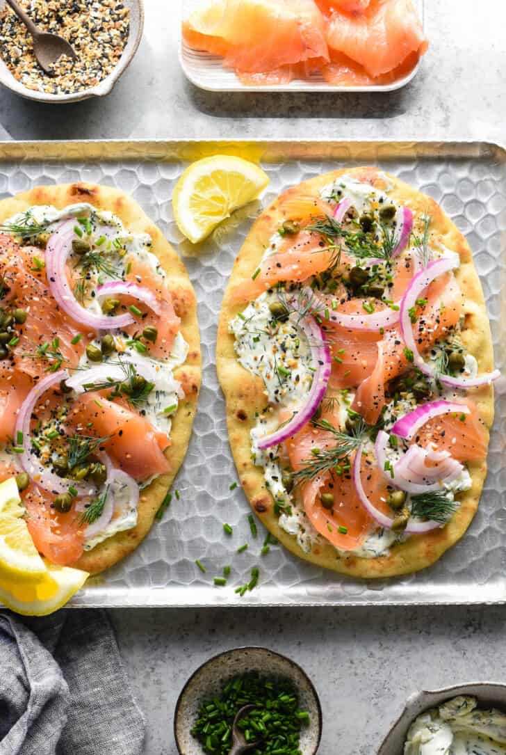 Flatbread pizzas on a metal textured tray made with naan bread, cream cheese, herbs, everything bagel seasoning, lox, capers and red onion.