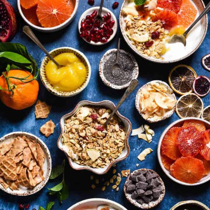 A yogurt bar set up on a blue background, with bowls of toppings including granola, waffle cone chunks, lemon curd, fruit, coconut, oranges and chocolate chunks.