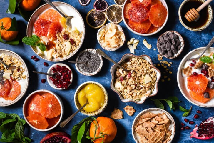 A variety of yogurt toppings set up on a blue background, including granola, waffle cone chunks, lemon curd, fruit, coconut, oranges and chocolate chunks.