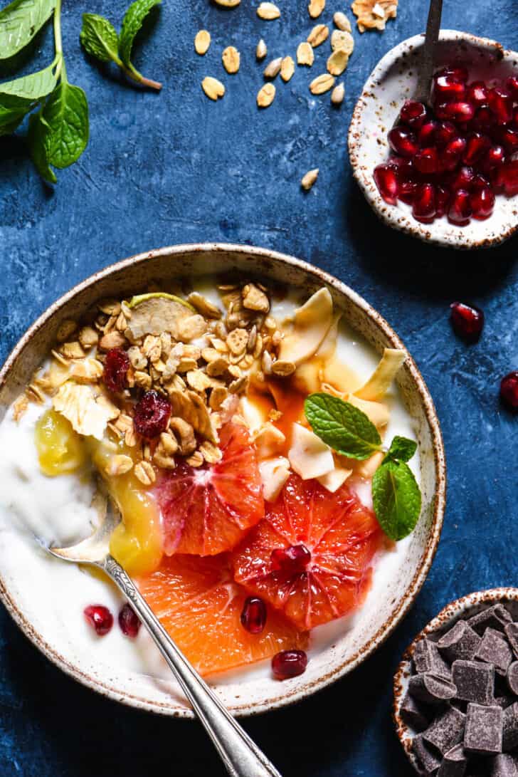 A textured bowl filled with yoghurt and topped with sliced oranges, granola, lemon curd, mint and  pomegranate seeds.