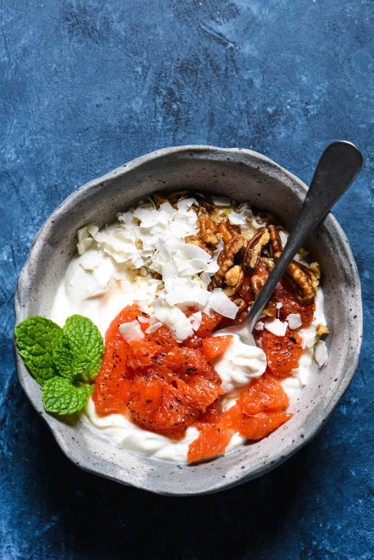 A bowl of yoghurt topped with bruleed grapefuit pieces, coconut flakes, pecans and fresh mint.