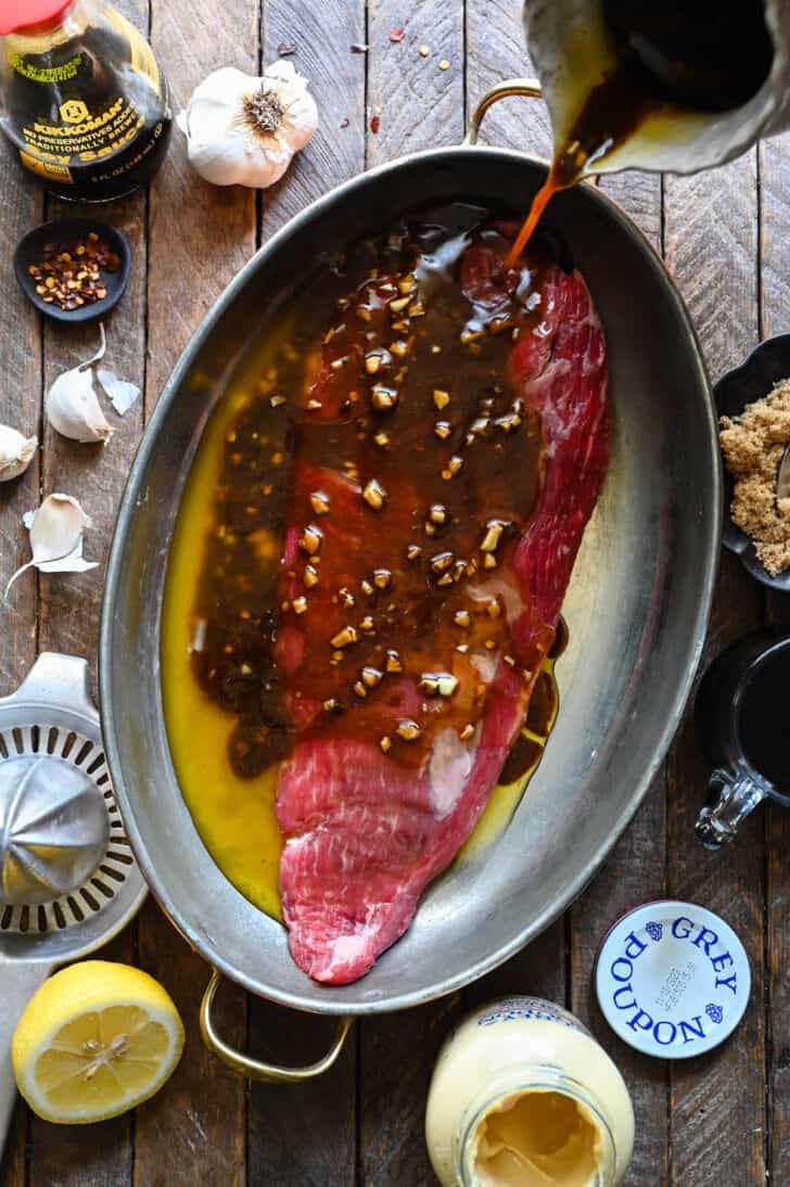 A piece of beef in a oval pan, with a hand pouring balsamic steak marinade with flecks of garlic in it over the meat.