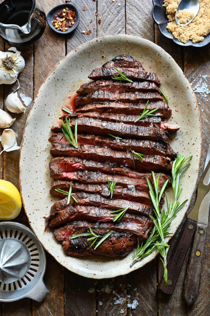 A cooked, sliced flank steak that has been marinated in balsamic steak marinade, on a speckled platter, with rosemary garnishing the steak.