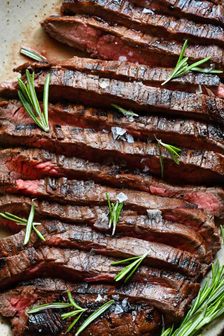 A cooked, sliced flank steak that has been marinated in balsamic steak marinade, on a speckled platter, with rosemary garnishing the steak.