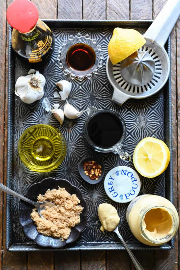 Ingredients needed for a marinade for the steak, laid out on a textured baking pan, including vinegar, oil, brown sugar, mustard, lemon, garlic, soy sauce and spices.