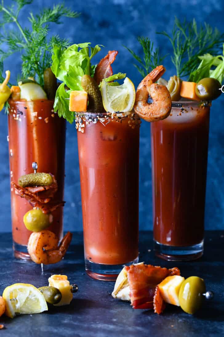 Three tall glasses filled with red juice, garnished with Bloody Mary skewers made with shrimp, bacon, cheese, pickles, artichokes and olives.