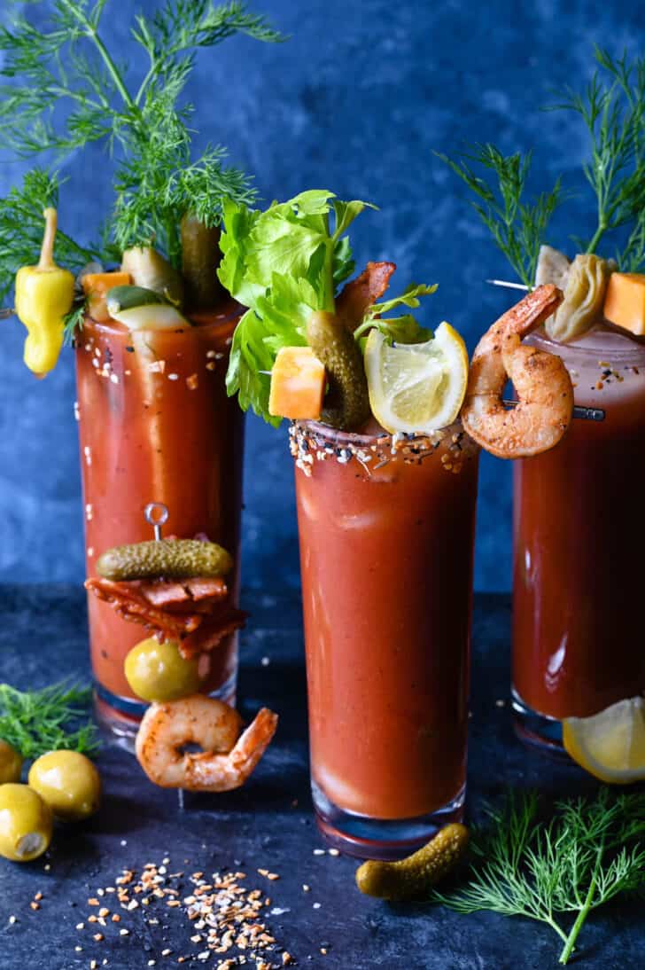 Three tall glasses of tomato juice with skewers that have garnishes for bloody mary, including shrimp, lemon wedges, cheese and pickles.