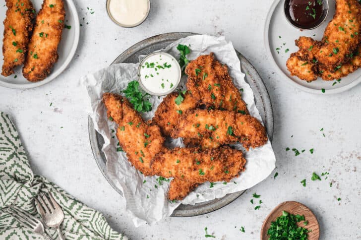 Fried buttermilk chicken tenders on a parchment-lined plate with a side of ranch dressing, sprinkled with parsley.