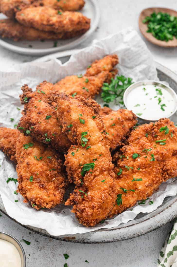 Fried buttermilk chicken tenders on a parchment-lined plate with a side of ranch dressing, sprinkled with parsley.