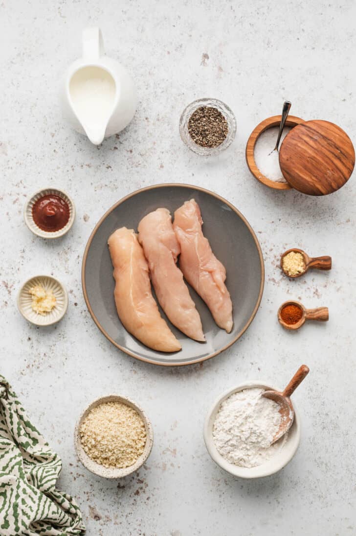 Ingredients laid out on a light surface, including poultry slices, seasonings, garlic, hot sauce, flour and breadcrumbs.