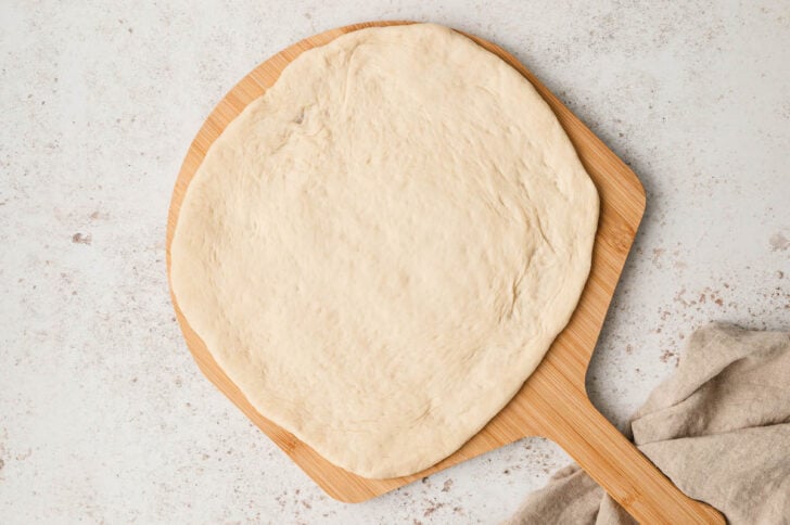 A wooden dough peel with a round of raw dough on it.