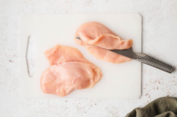 A knife slicing chicken breasts in half on a plastic cutting board.