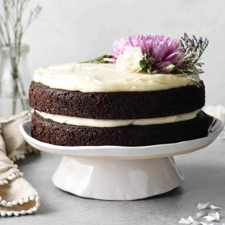 A two layer chocolate cake with cream cheese frosting on a white cake stand, decorated with pink and white flowers and greenery.