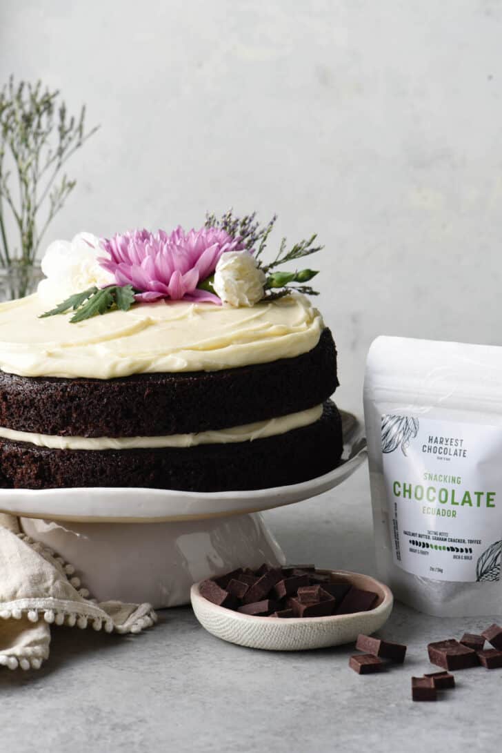 A black and white later cake on a white ceramic cake stand with a bag of Harvest Chocolate snacking chocolate alongside the cake.