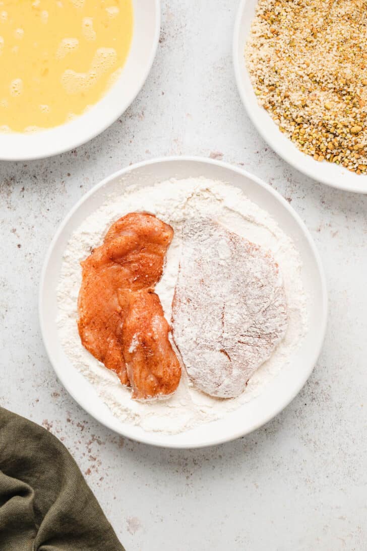 Seasoned chicken breasts being coated with flour in a bowl.