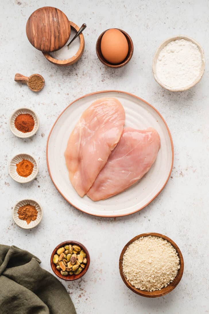 A white surface with the ingredients needed for pistachio chicken, including chicken breasts, flour, breadcrumbs, spices, an egg and pistachios.