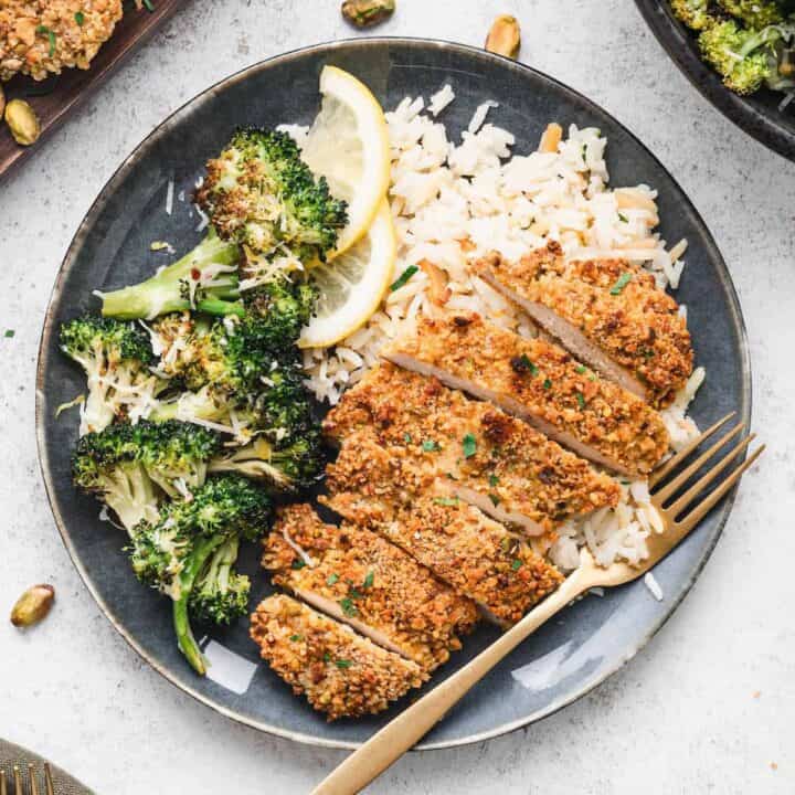 A blue plate topped with rice, roasted broccoli and sliced pistachio crusted chicken.