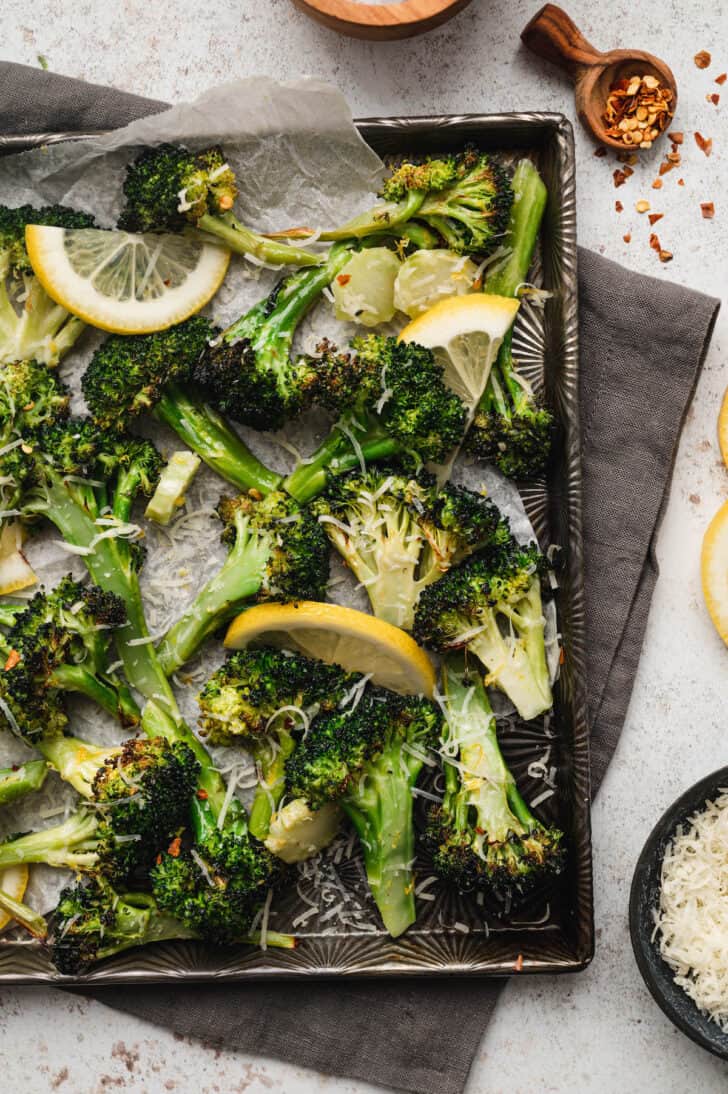 Roasted broccoli with lemon slices, shredded cheese and red pepper flakes on a textured baking pan.
