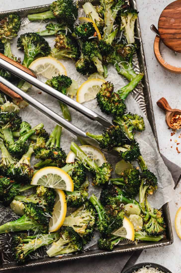 Textured sheet pan with parchment paper, topped with roasted broccoli with lemon slices, Parmesan cheese and red pepper flakes, with tons removing some of the broccoli.