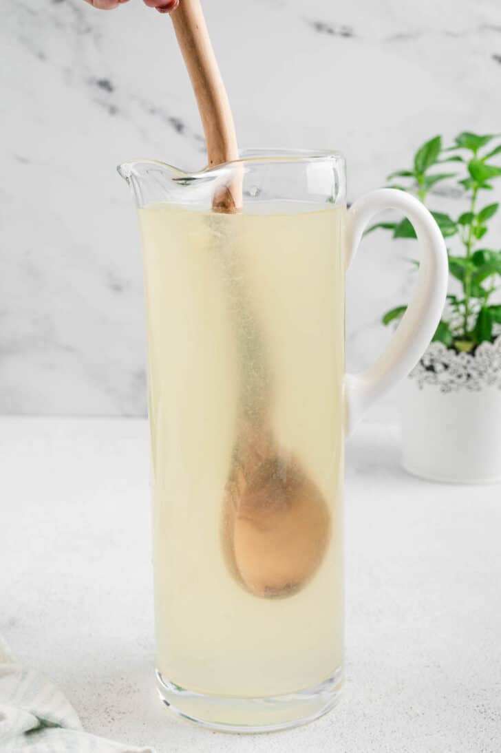 A tall glass pitcher filled with a light yellow liquid, being stirred with a wooden spoon.