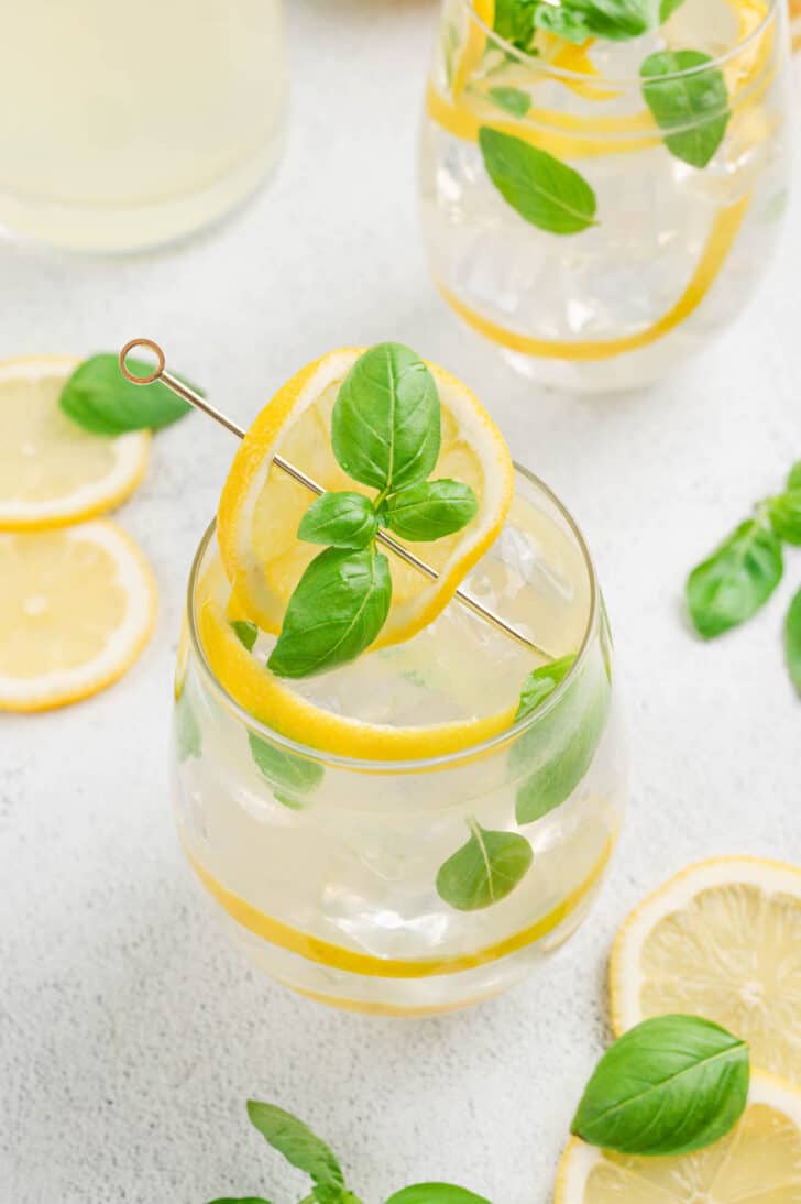 A glass filled with ice and basil lemonade, garnished with lemon peels and fresh basil leaves.
