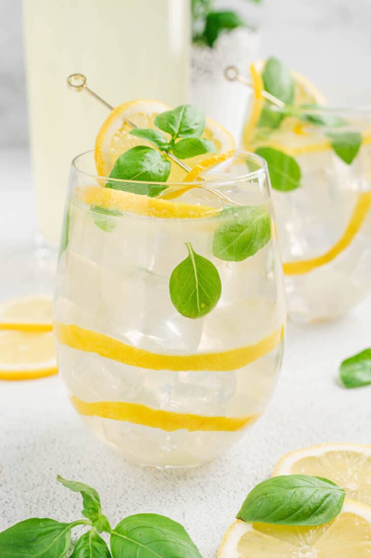 A glass filled with ice and a light yellow liquid, garnished with lemon peels and fresh herb leaves.