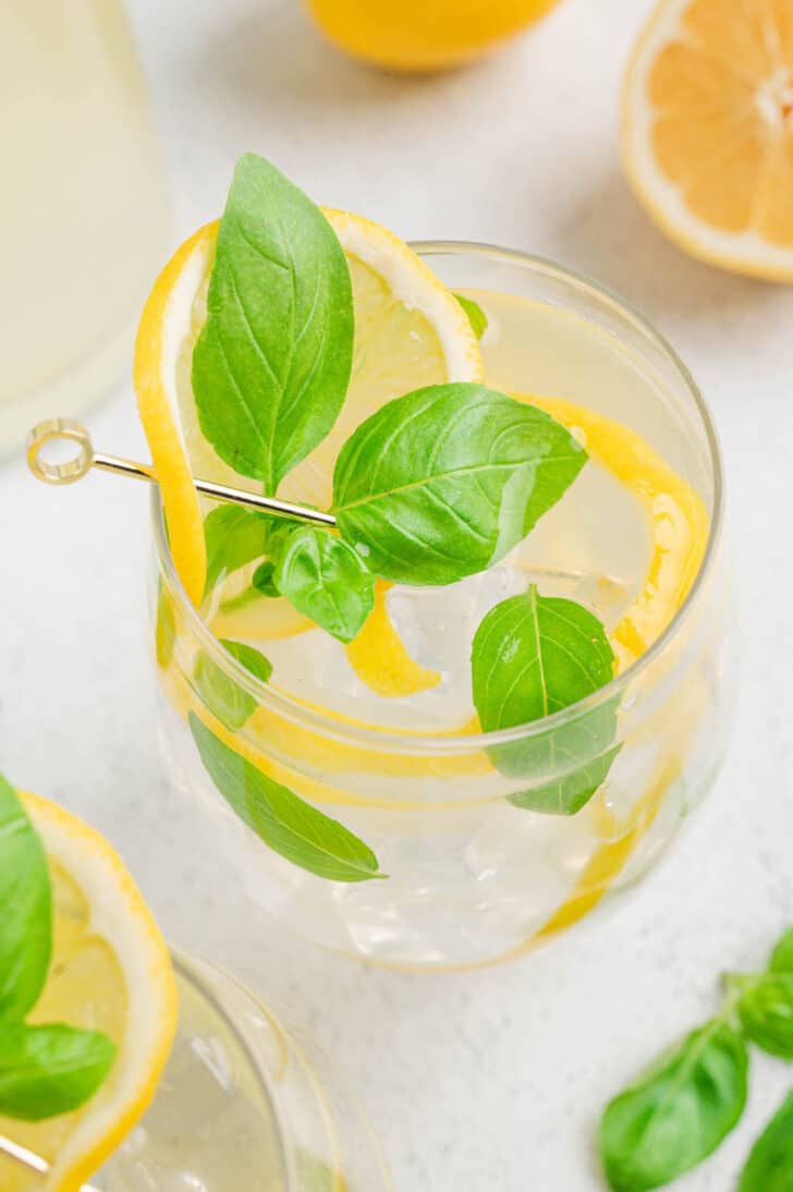 A glass filled with ice and basil lemonade, garnished with lemon peels and fresh basil leaves.