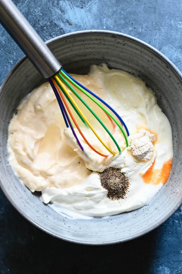 A gray bowl filled with mayonnaise, sour cream, spices and hot sauce, with a rainbow whisk getting ready to stir everything up.