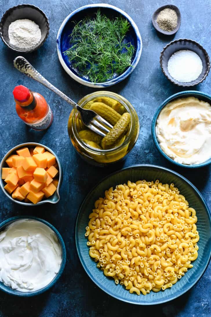 Ingredients laid out on a blue surface including macaroni noodles, cheese, mayonnaise, dill, hot sauce and pickled cucumbers.