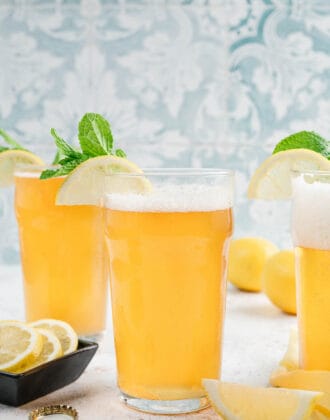 Glasses of lemon shandy garnished with lemon wedges and fresh mint in front of a patterned blue background.