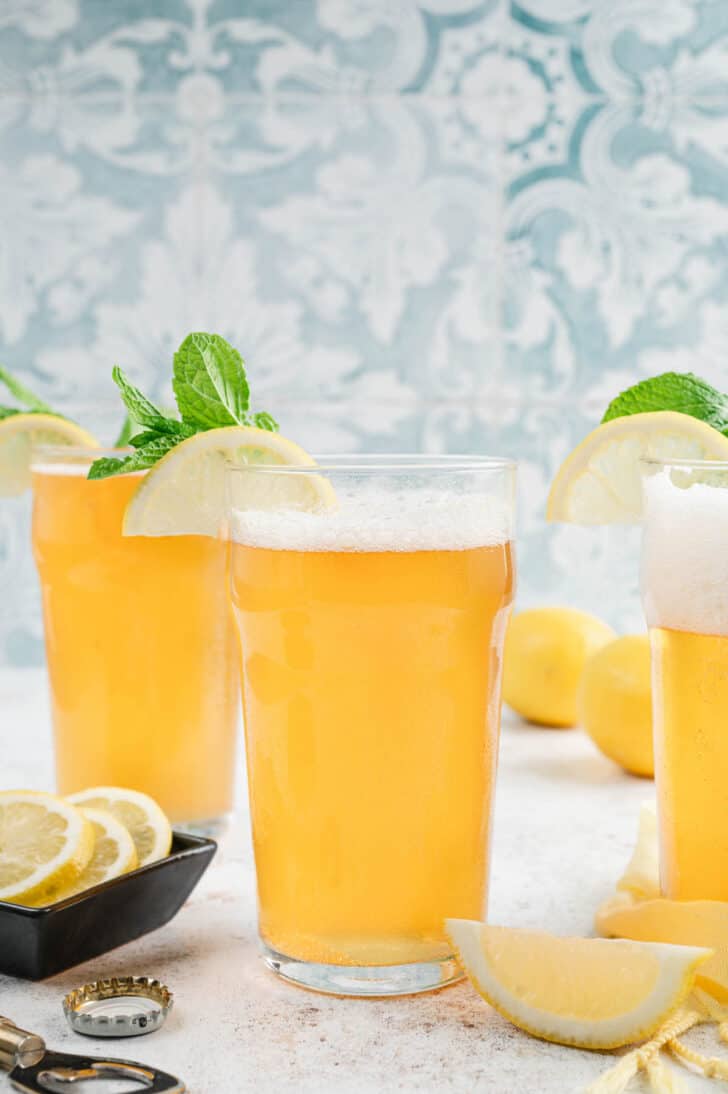 Glasses of lemon shandy garnished with lemon wedges and fresh mint in front of a patterned blue background.