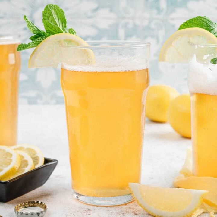 Glasses of lemon shandy garnished with lemon wedges and fresh mint in front of a patterned blue background.