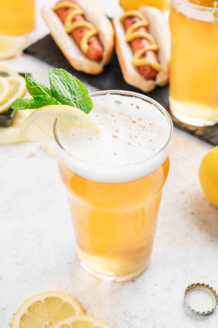A pint glass filled with beer and lemonade, garnished with mint and lemon, with hot dogs in the background.