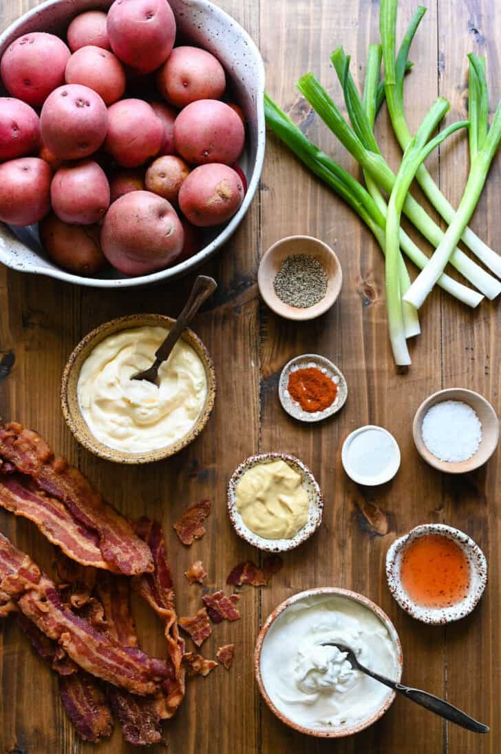 Ingredients laid out on a wooden surface, including red spuds, green onions, mayonnaise, sour cream, mustard, spices and bacon.
