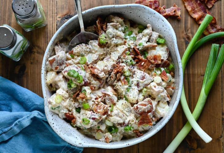 A bowl of bacon potato salad on a wooden surface with green onions, bacon, salt and pepper shakers and a blue napkin decorating the scene.