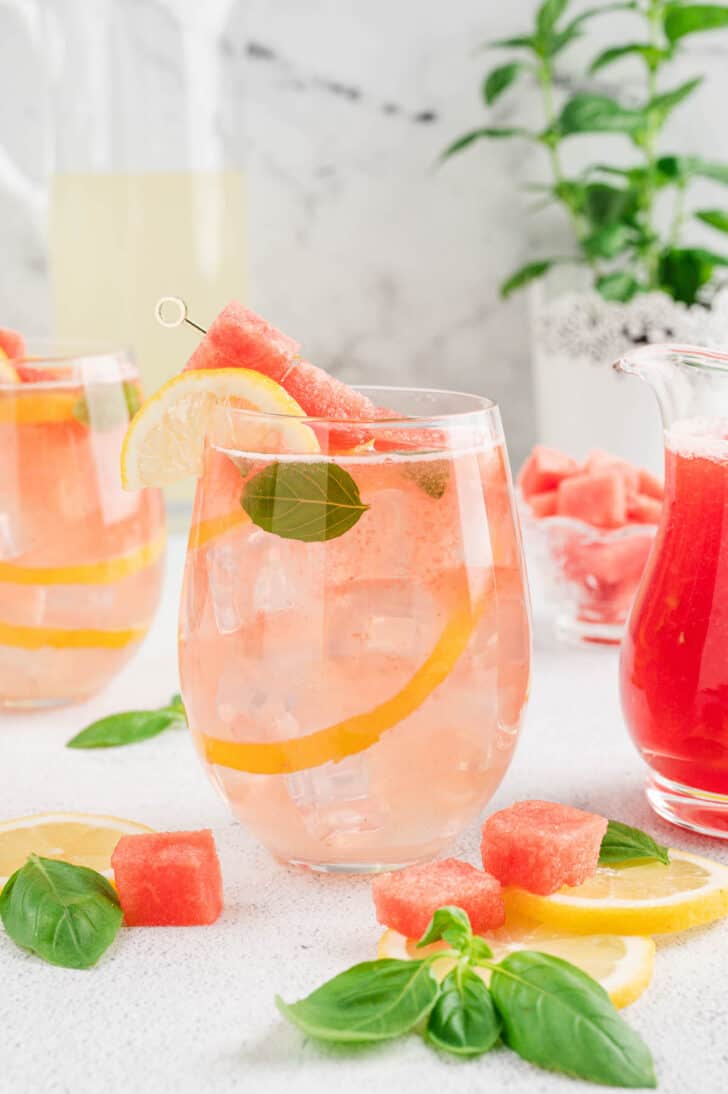 A glass of watermelon basil lemonade garnished with a skewer of cubed watermelon, a lemon slice, and fresh basil leaves.