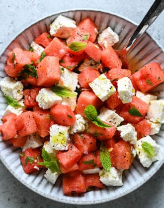 A textured gray bowl filled with watermelon feta mint salad, with a spoon digging into it.