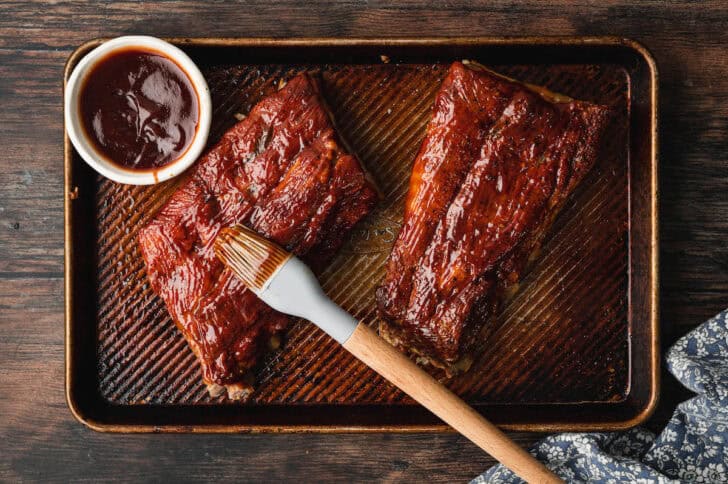 Two racks of bone-in pork being brushed with barbecue sauce.