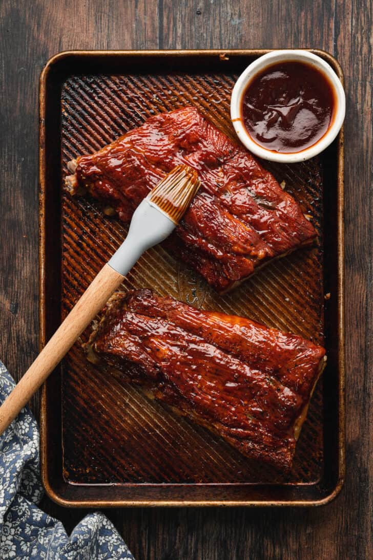 Bone in pork meat on a textured baking pan being brushed with barbecue sauce.