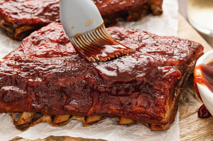 A rack of baked baby back ribs being brushed with barbecue sauce.