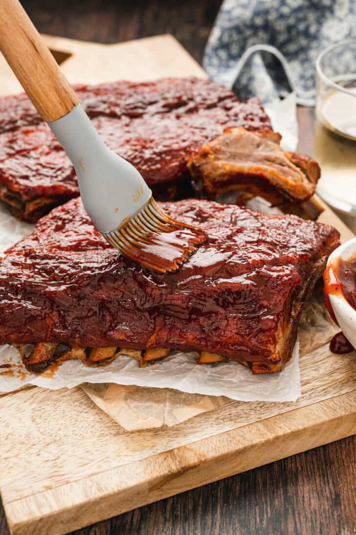 Oven baked baby back ribs being brushed with barbecue sauce, on a wooden cutting board and butcher paper.