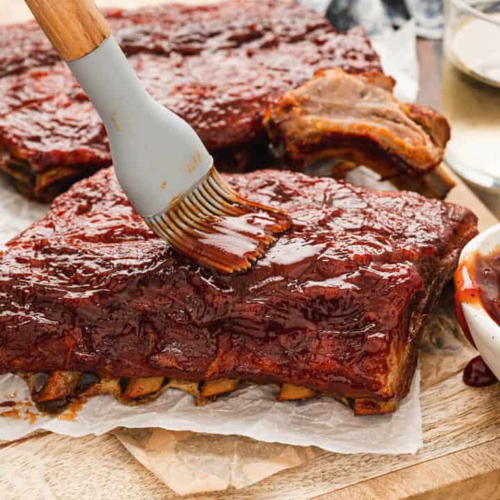 Oven baked baby back ribs being brushed with barbecue sauce, on a wooden cutting board and butcher paper.